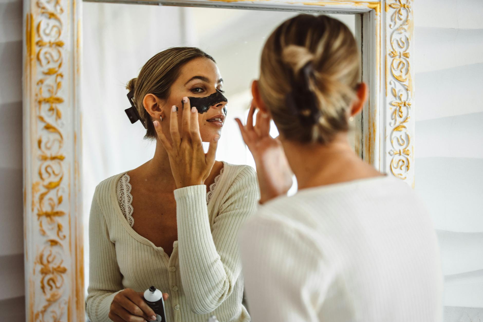 Najczęstsze błędy przy stosowaniu masek do włosów i jak wybierać maskę do aktualnych potrzeb fryzury Kobieta nakłada czarną maseczkę na twarz, patrząc w lustro w łazience