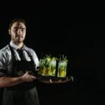 Uśmiechnięty barman w uniformie trzyma tacę z cytrynowymi drinkami