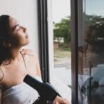 Kobieta suszy włosy suszarką przy otwartym oknie w domu